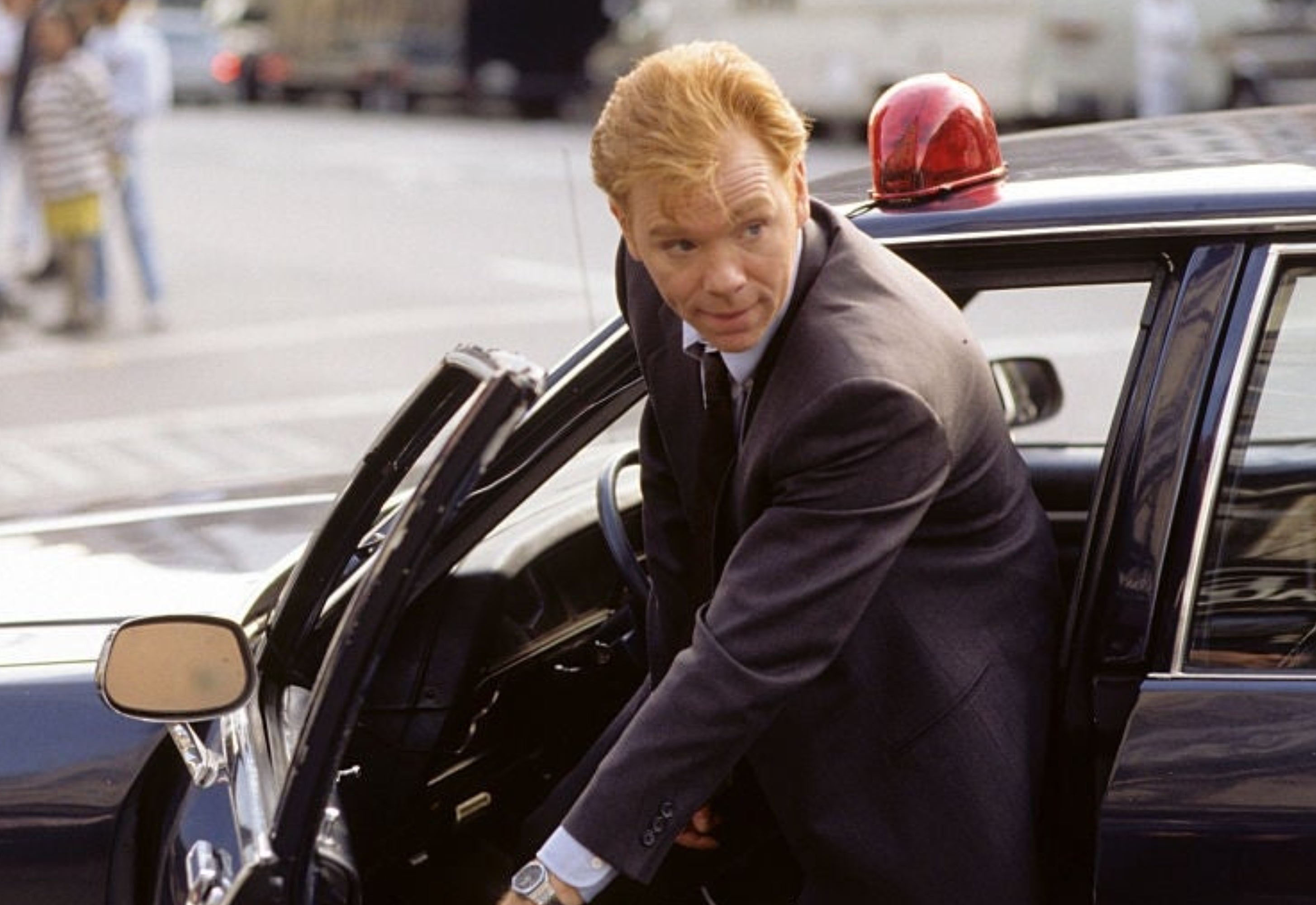 A man wearing a suit is stepping out of a police car, with people and a street scene in the background
