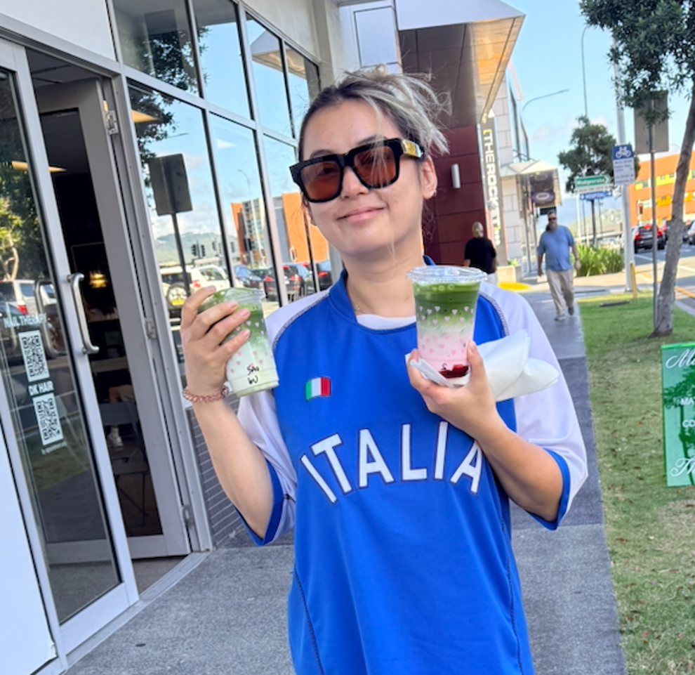 Person outdoors holding a drink and food, wearing large sunglasses, a casual oversized sports jersey, and sandals
