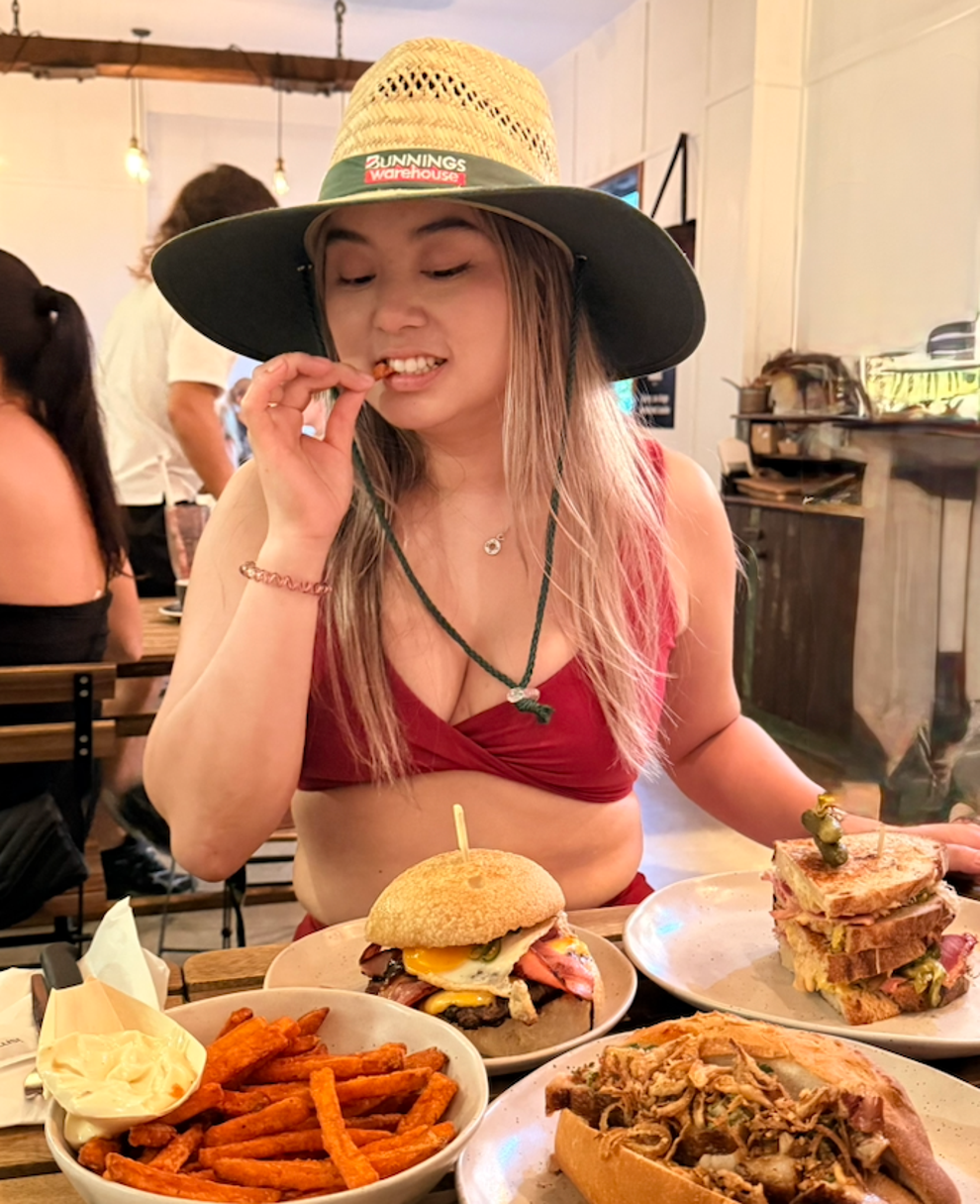 Person in a casual hat and bikini top enjoys a meal at a restaurant table filled with burgers and sweet potato fries
