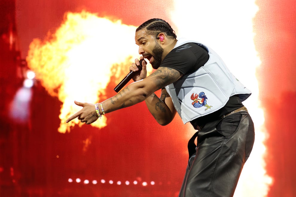 Performer on stage with a microphone, wearing a light vest and dark pants, gestures energetically in front of a bright fire effect