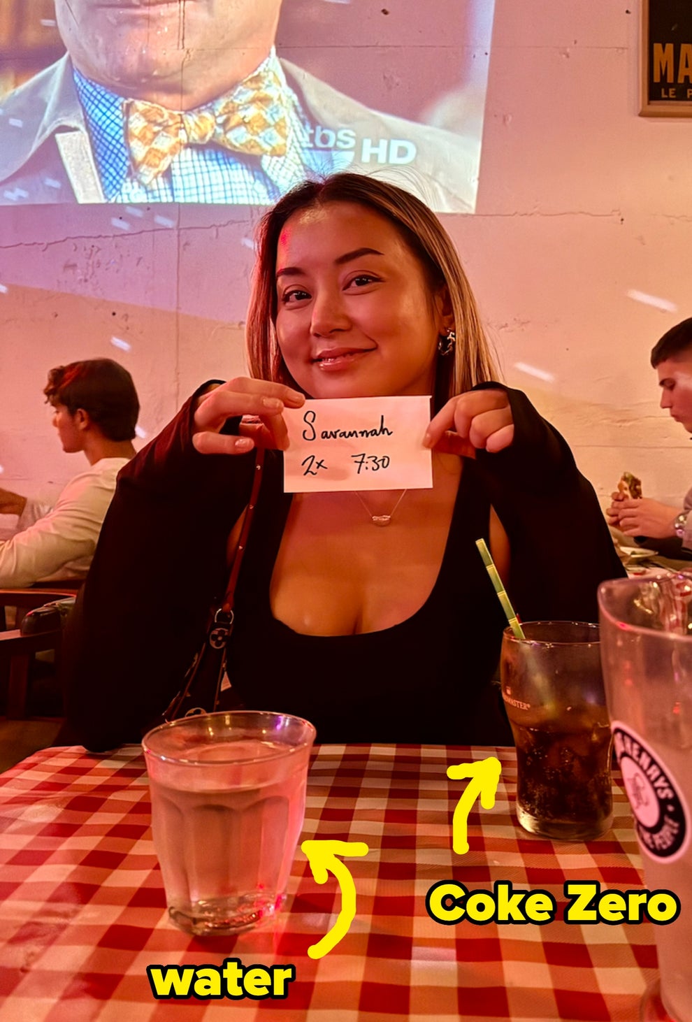Woman at a restaurant holding a reservation slip with the name "Savannah," date "4x," and time "7:30." Two people are seated in the background