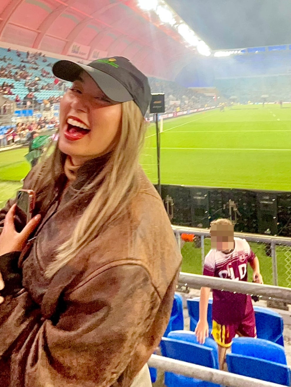A person in a casual jacket and cap smiles joyfully in a sports stadium, with bright lights and a grassy field in the background