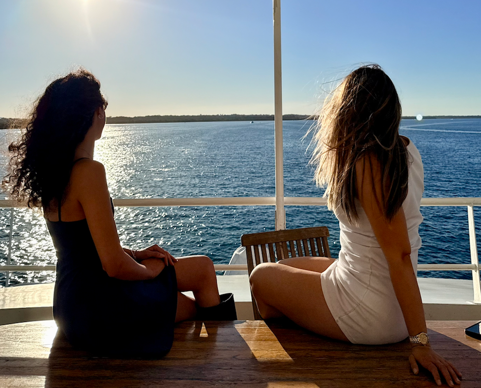 Two people sitting on a boat with an ocean view, under a clear sky. One is wearing a white sleeveless dress and the other a dark outfit