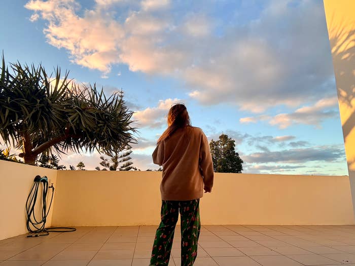 Person stands on a terrace in pajamas, looking at the cloudy sky, near a potted tree and garden hose