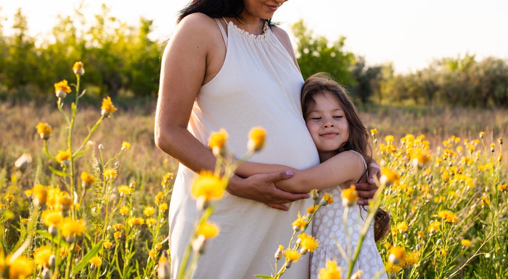 Pregnant woman in a field with yellow flowers, holding hands with a young girl. They both wear light dresses and share a tender moment