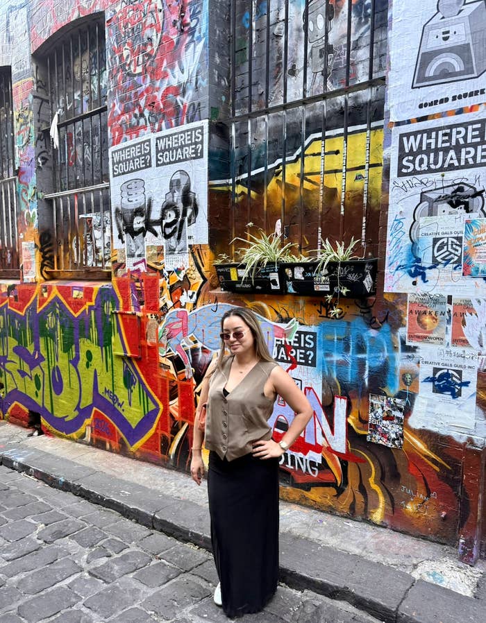 Person standing confidently in front of a graffiti-covered wall, wearing sunglasses, a sleeveless top, and a long skirt