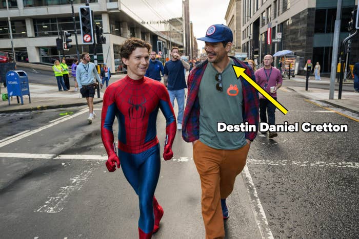 Person in Spider-Man costume walking with another person in casual attire on a city street, smiling and engaged in conversation