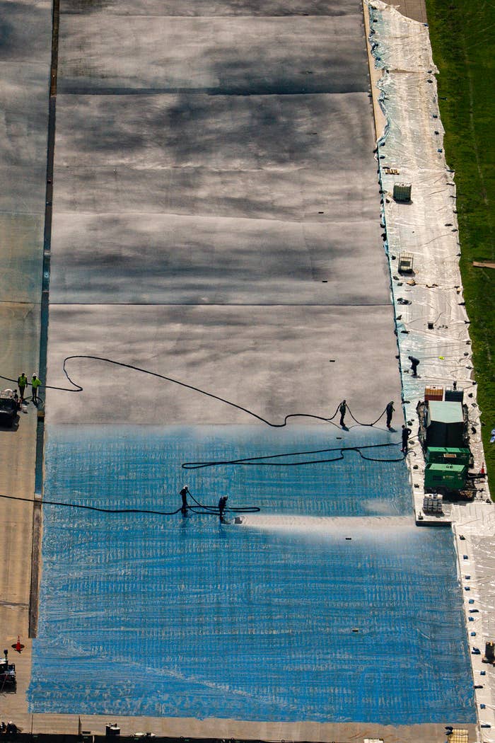 Workers applying a blue coating on a large surface, using hoses and machinery, viewed from above
