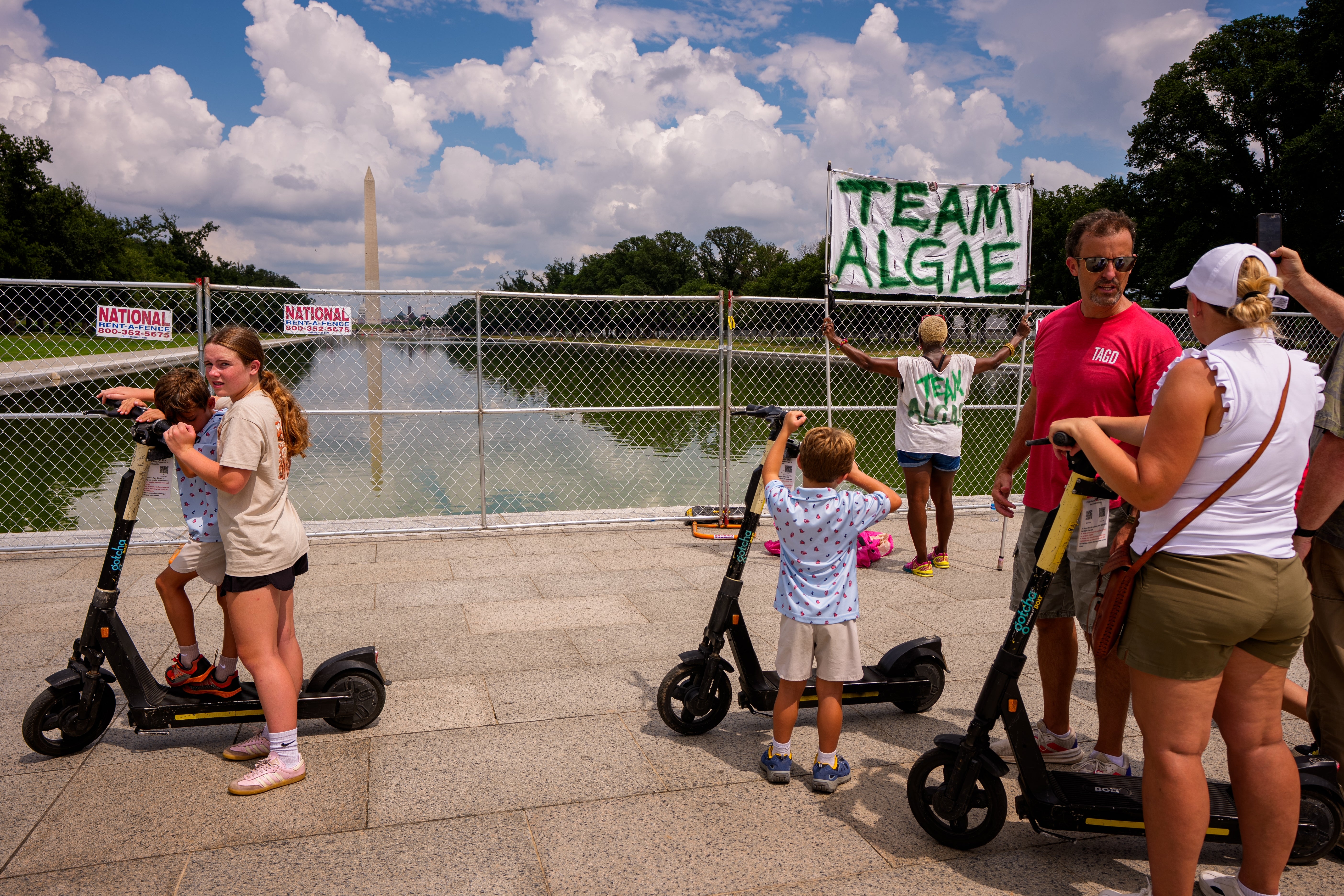 Folk på scootere ved Washington-monumentet; noen holder en "Team Algae" skilt nær et inngjerdet reflekterende basseng