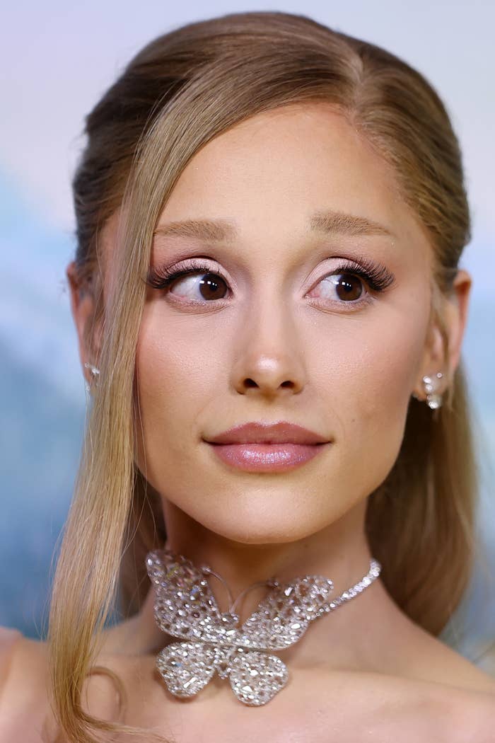 Person with long hair and elegant makeup looks to the side, wearing a detailed butterfly necklace