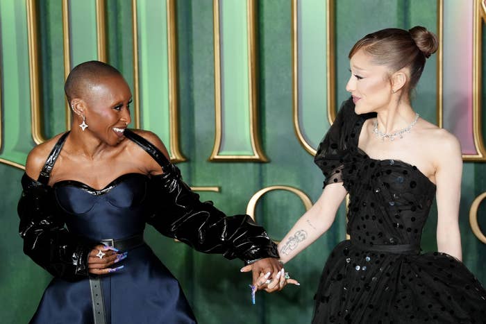 Two individuals in elegant black dresses hold hands on a red carpet, sharing a moment of joy