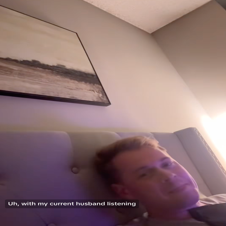 Person lying on a bed, looking sideways. Text overlay: "Uh, with my current husband listening."