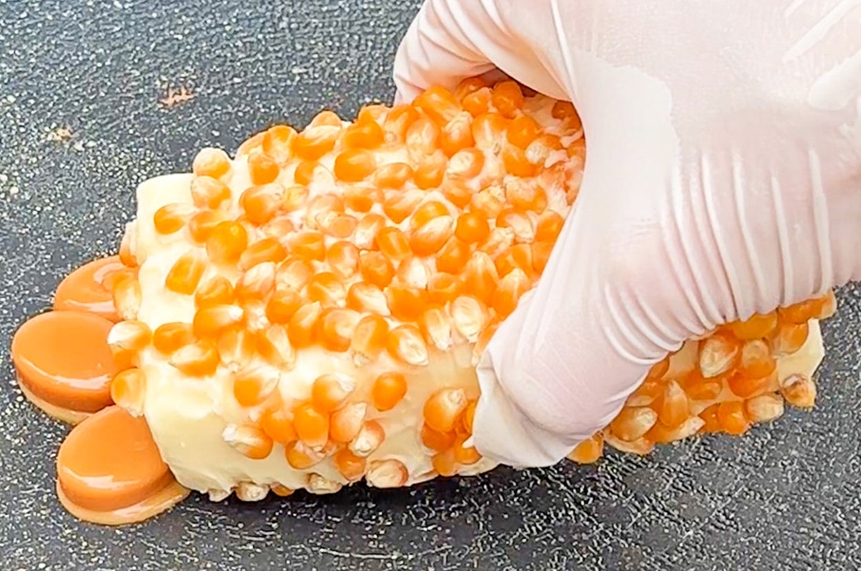 ポップコーンの種をまぶしたクリーム色のアイスクリームバーを手袋をした手が持っている。