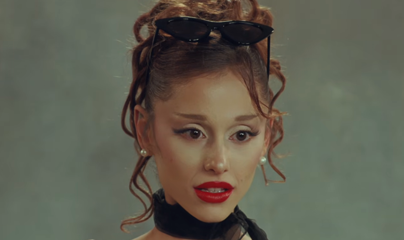 Person with curly hair and sunglasses resting on head, wearing pearl earrings and a black neck scarf