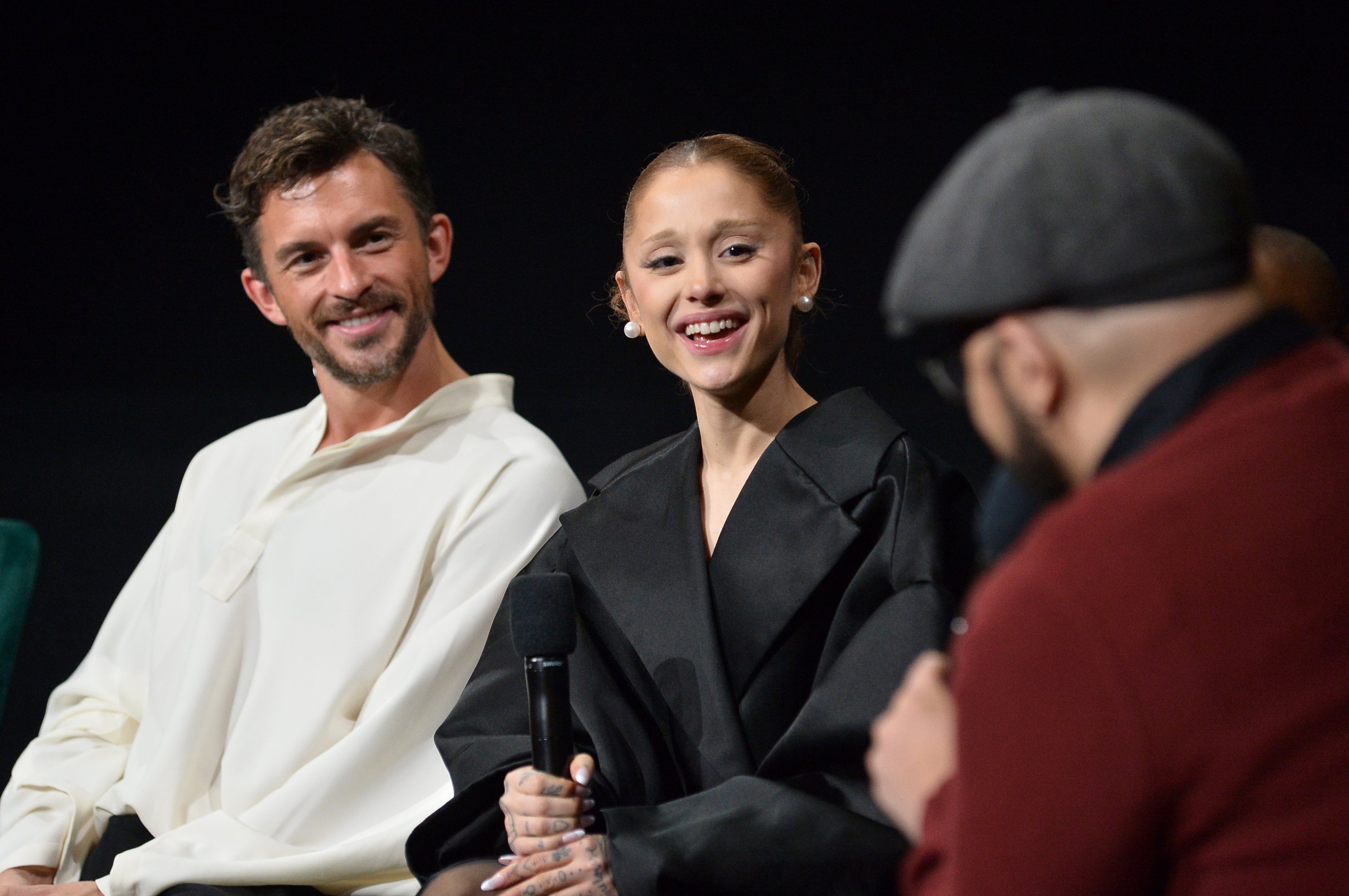 Two people in stylish attire sit on a panel engaging in conversation, holding microphones and smiling at another person speaking
