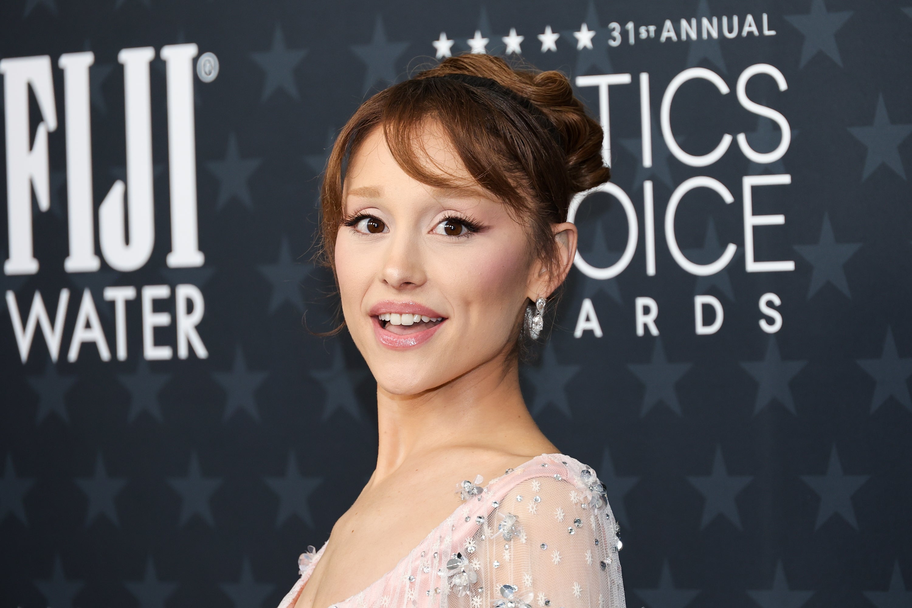 Person smiling at an award event, wearing an elegant dress with floral embellishments, hair styled in an updo. Background has awards branding