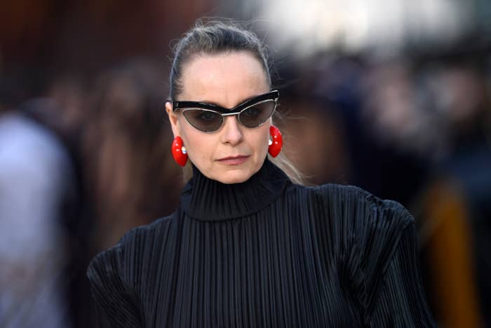 A person wearing a high-neck pleated black outfit, cat-eye sunglasses, and bold red earrings at a celebrity event