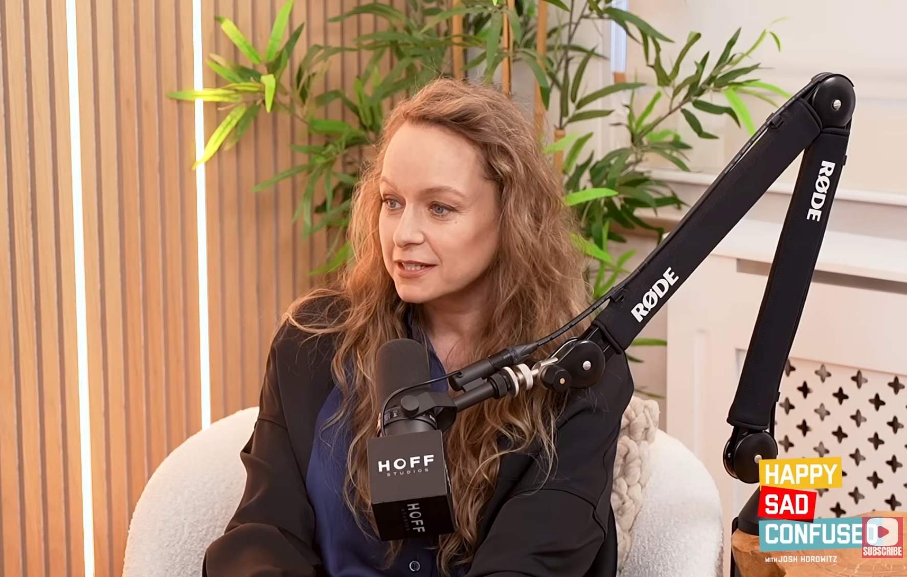 A person with long hair is speaking into a podcast microphone in a studio setting with plants in the background