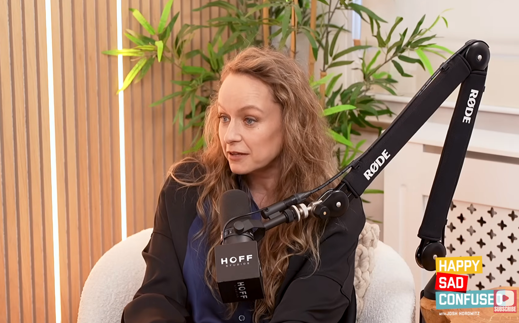 A person is speaking into a microphone on a podcast set, with plants in the background and soundproofing panels visible
