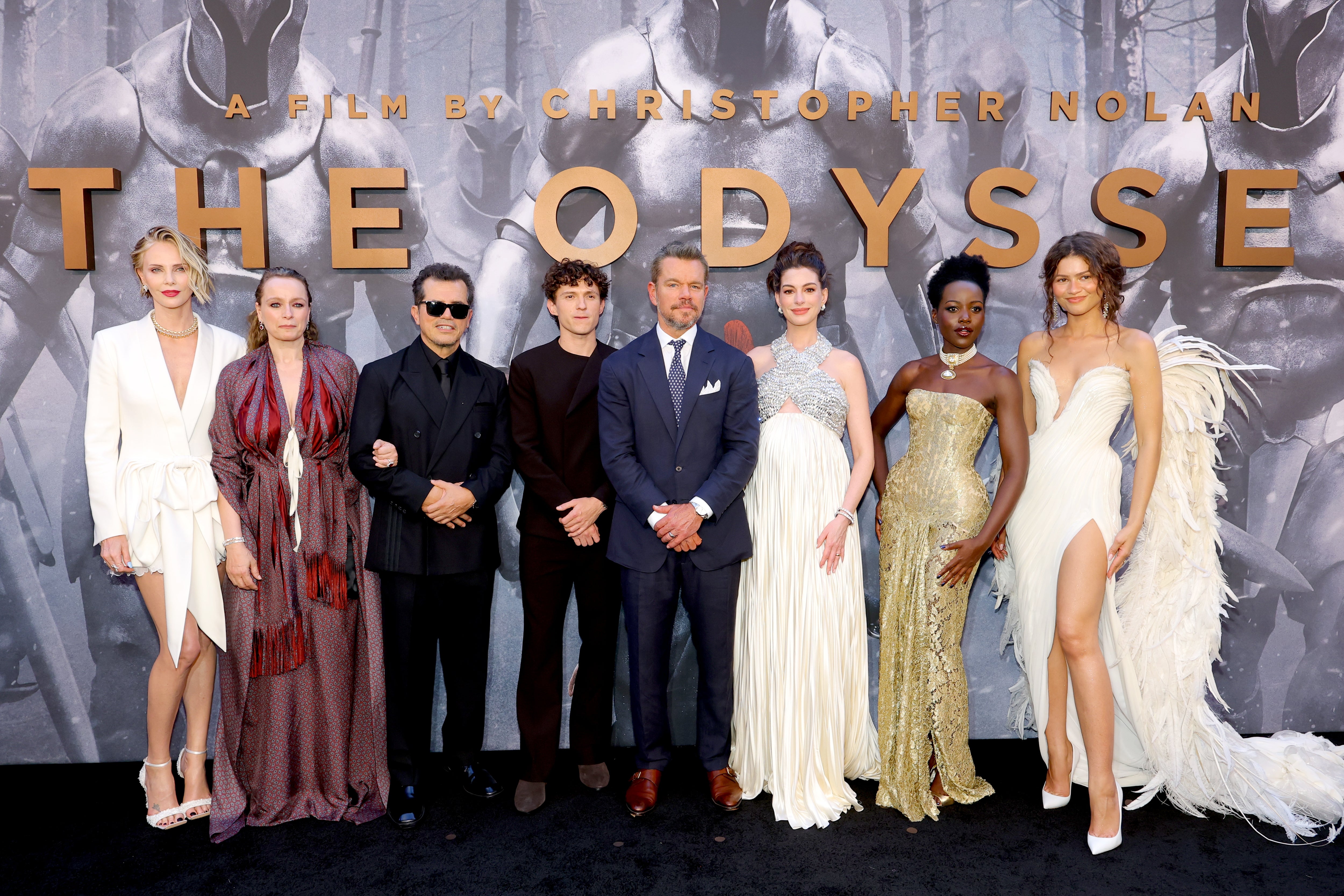 Group of actors in formal wear at a movie premiere, standing in front of a backdrop with "The Odyssey" text