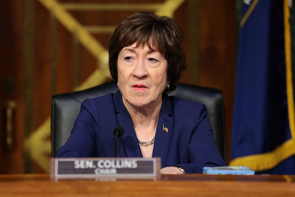 Susan Collins seated at a desk in a formal setting, wearing a blazer, appearing engaged in a discussion or hearing