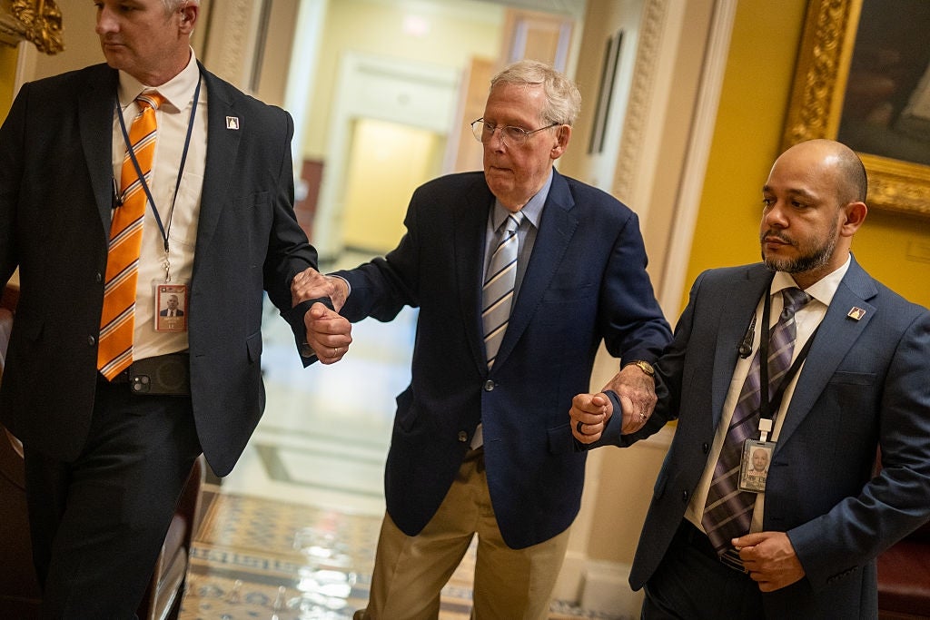 Mitch McConnell in a suit, assisted by two men, walks through a hallway