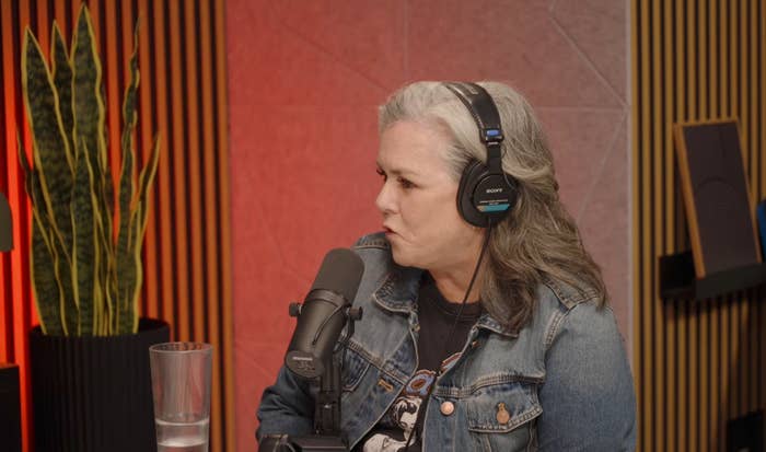 Person with headphones speaking into a microphone, seated in a podcast studio. A plant is visible in the background