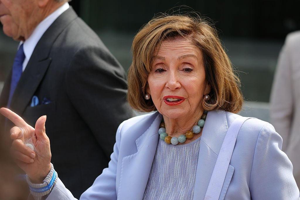 Nancy Pelosi in a formal suit and necklace speaks while gesturing, captured in an outdoor setting