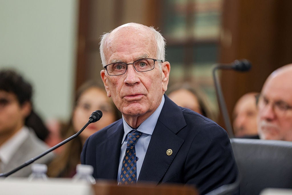 Peter Welch in glasses, wearing a suit and tie, speaks at a public event, surrounded by others in a formal setting