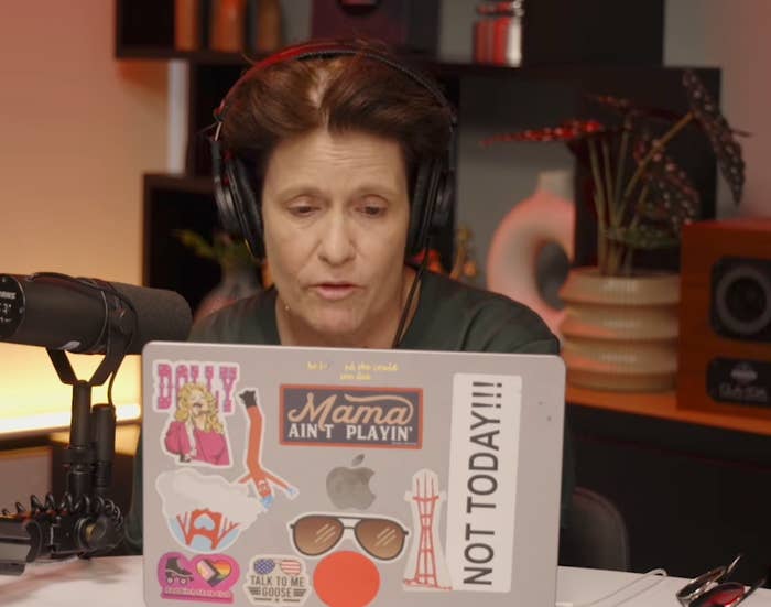 Person wearing headphones, speaking into a microphone, viewed from behind a laptop with various stickers, including text "Mama Ain't Playin'" and "NOT TODAY!!!"