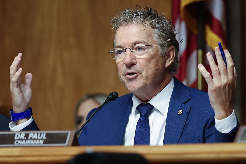 Rand Paul in a suit, gesturing with his hands, speaks at a podium with a "Dr. Paul, Chairman" nameplate during a formal event