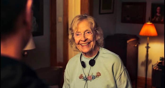 Elderly woman smiling indoors, wearing a floral sweatshirt and headphones around her neck