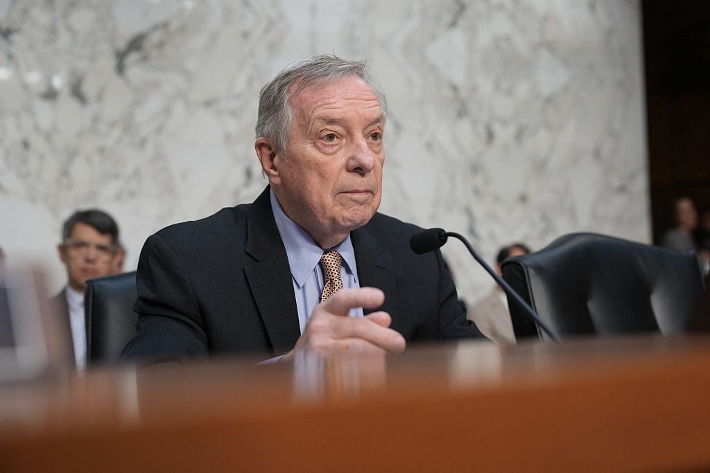 Dick Durbin in a suit speaks into a microphone at a formal indoor event, seated at a large table with others in the background