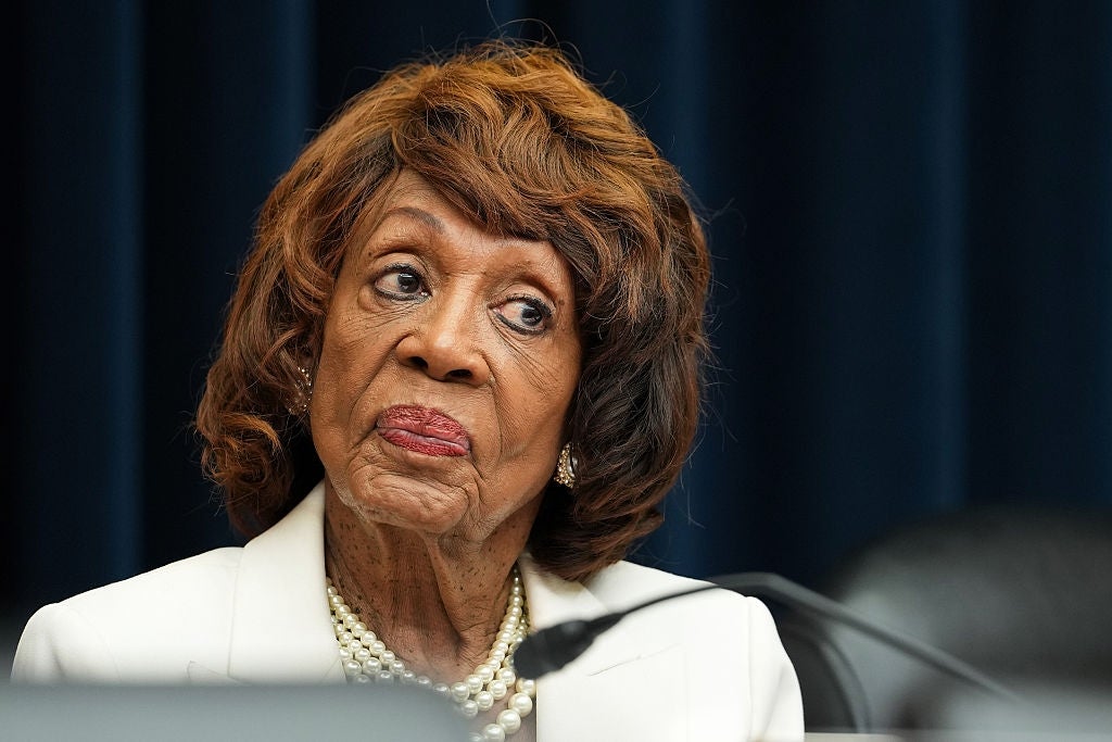 Maxine Waters wearing a white blazer and pearl necklace, sitting at a table, appears engaged in a serious discussion at a formal event