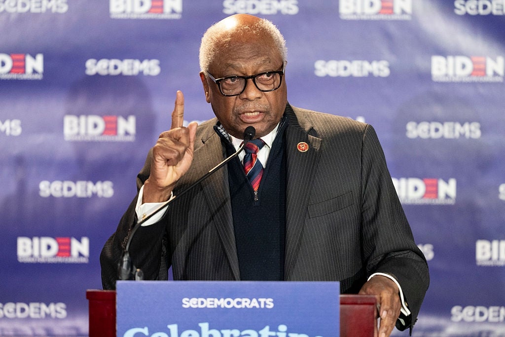 Jim Clyburn speaking at a podium with a "SCDemocrats" backdrop, gesturing and dressed in a suit and tie