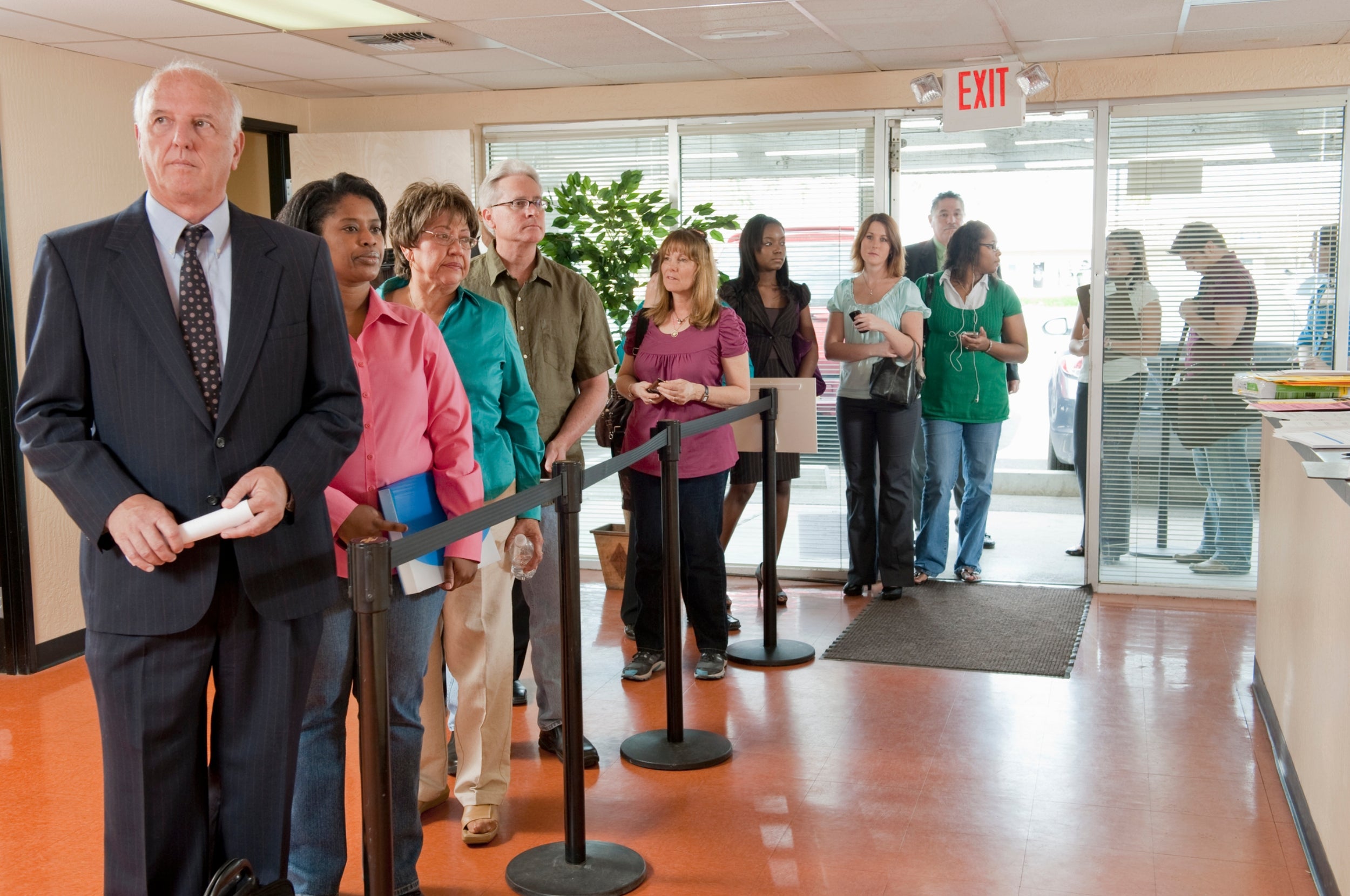 People of various ages and styles wait in line inside a building with glass doors and a plant