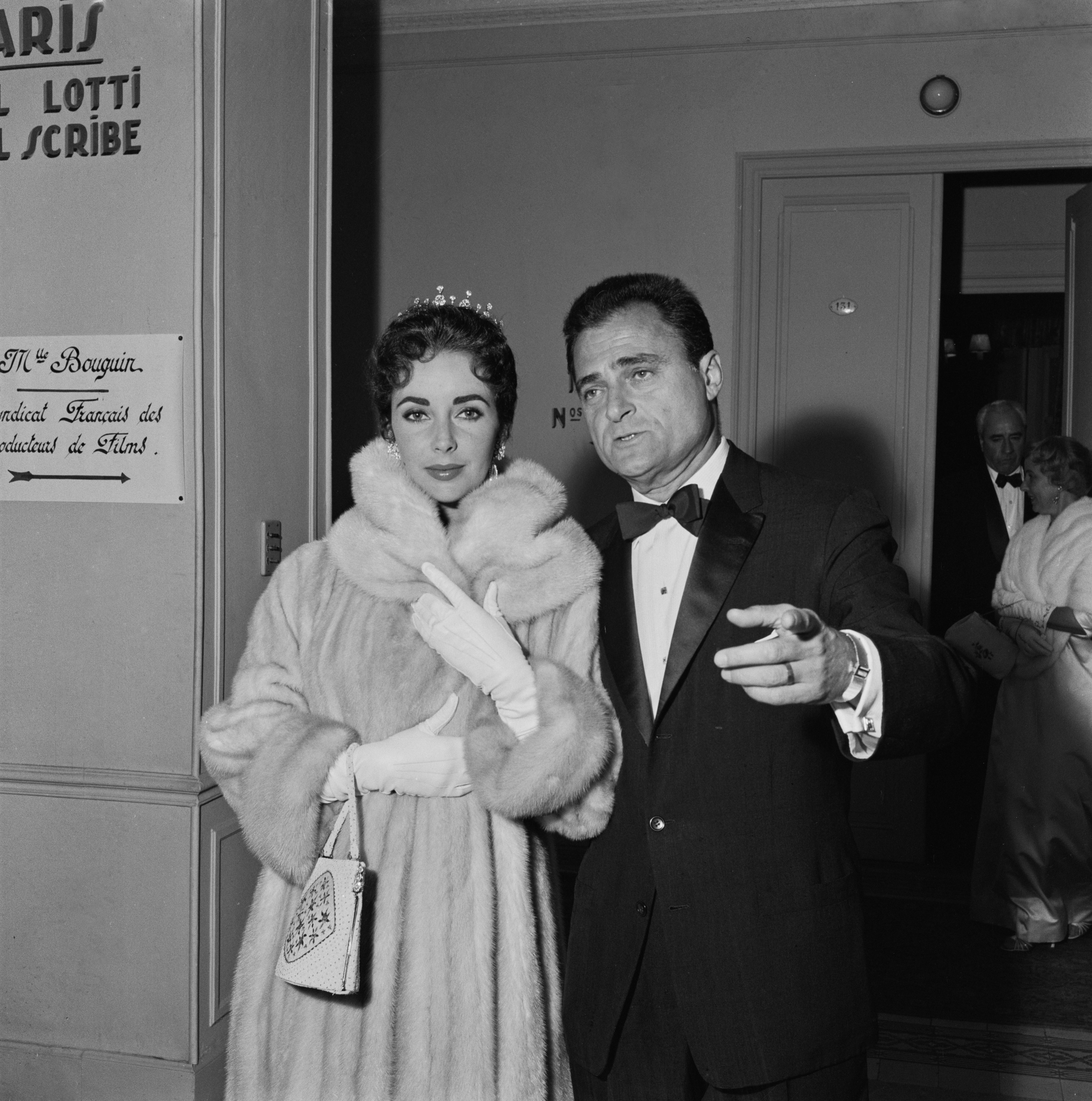 Elizabeth Taylor and Mike Todd in formal attire, one wearing a fur coat and gloves, stand in a classic setting, appearing poised and engaged