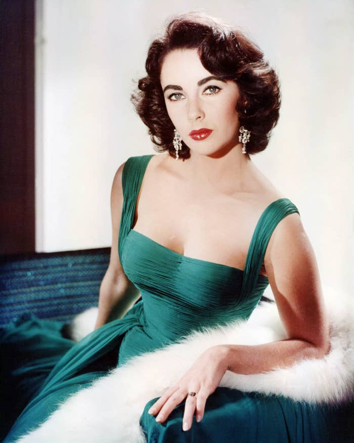 A person in an elegant, vintage-style gown poses indoors on a fur-covered seat, with short, styled hair and chandelier earrings