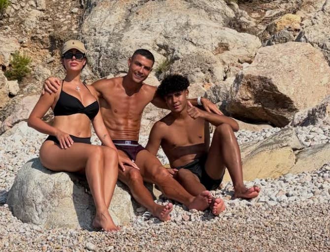 Three people sit on rocky beach, smiling. Left: woman in bikini and cap. Center: shirtless man. Right: young man in shorts giving thumbs-up