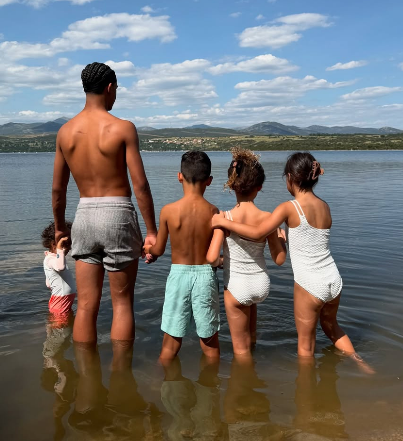 A family of five, wearing swimsuits, stand in shallow water at a lake, facing away, with scenic mountains and clouds in the background