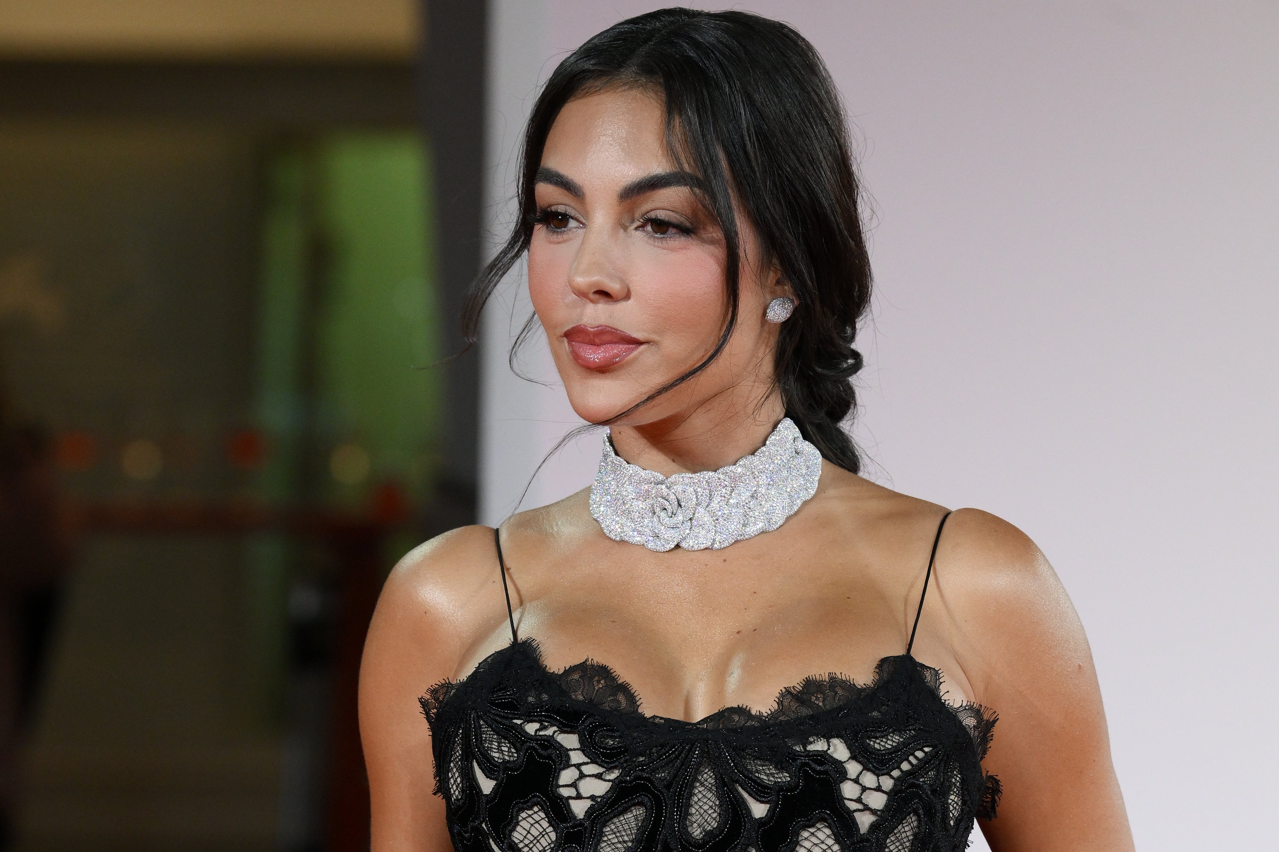 Person in elegant, lace-patterned dress with a sparkling choker, posing on a red carpet. Hair is styled in a neat updo