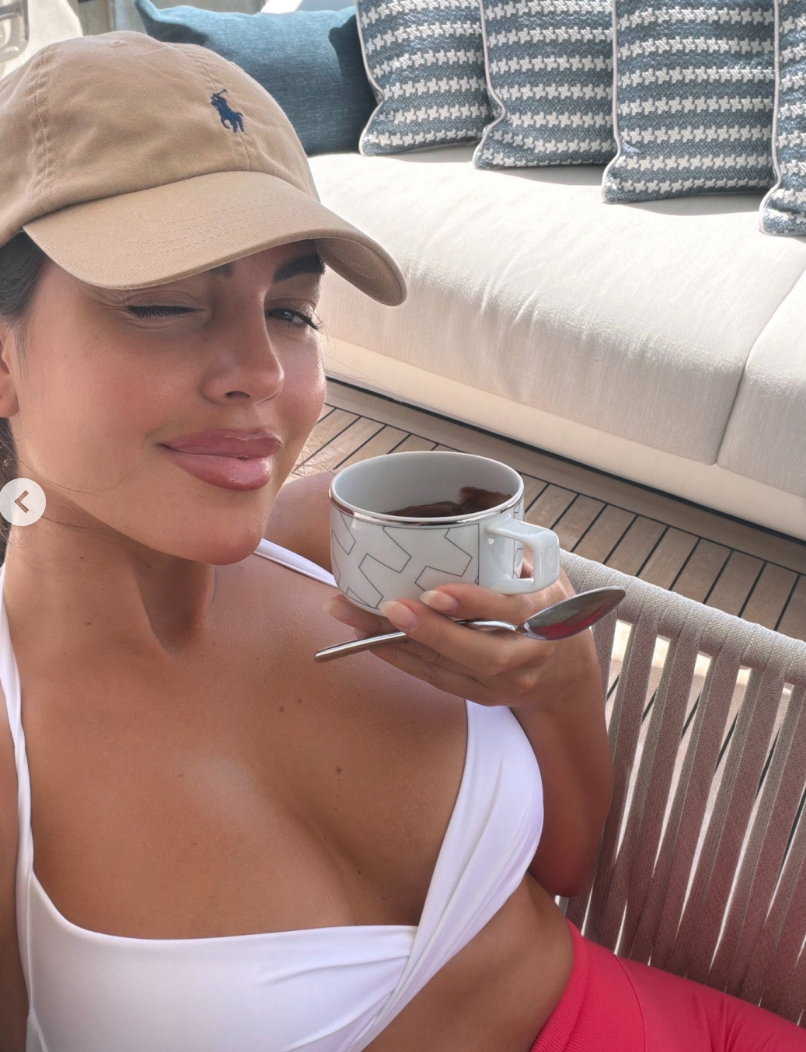 Person in a cap winks while holding a tea cup, sitting on a boat deck with houndstooth pillows in the background