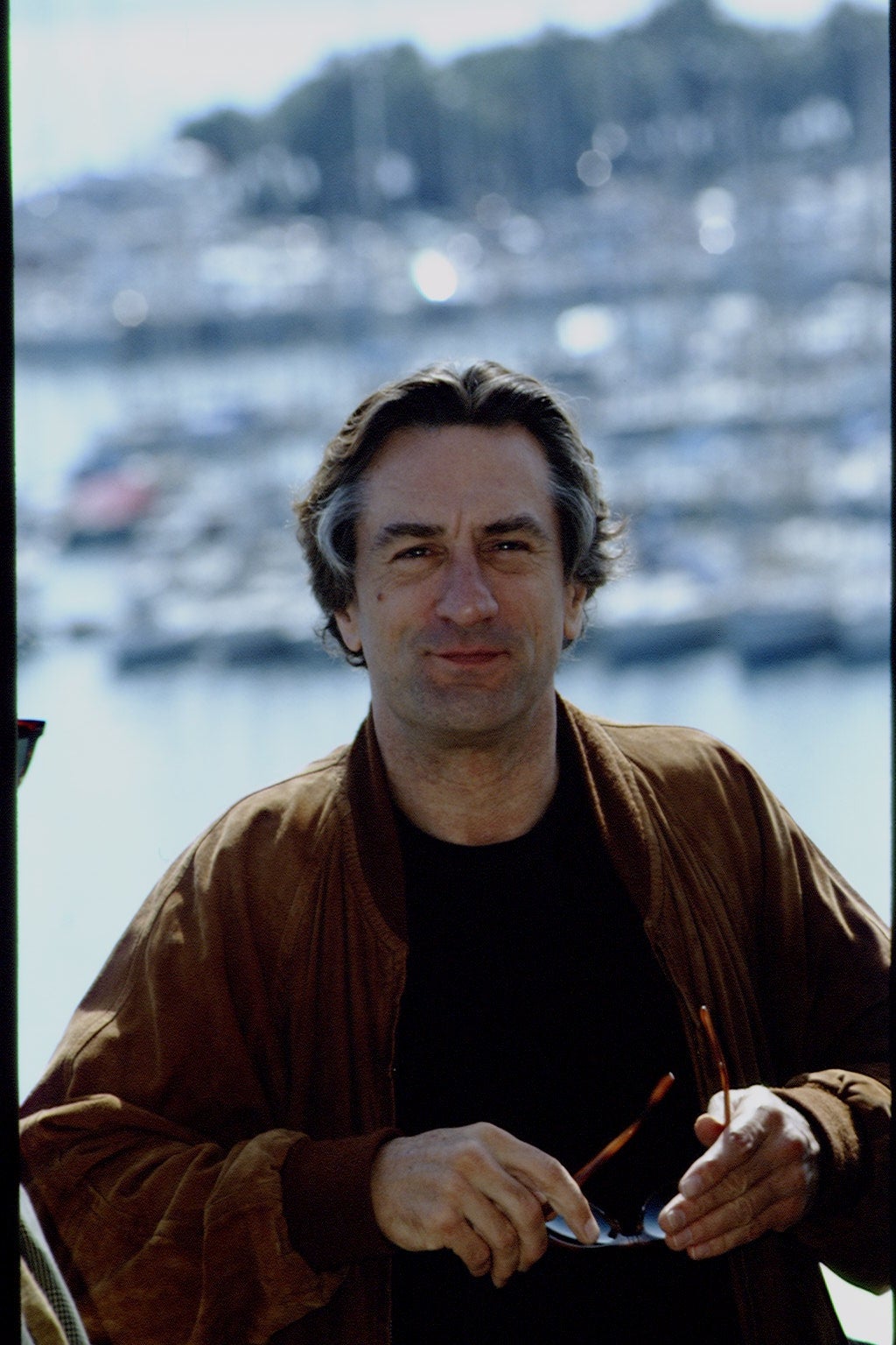 A person standing outdoors, holding sunglasses, with a marina backdrop