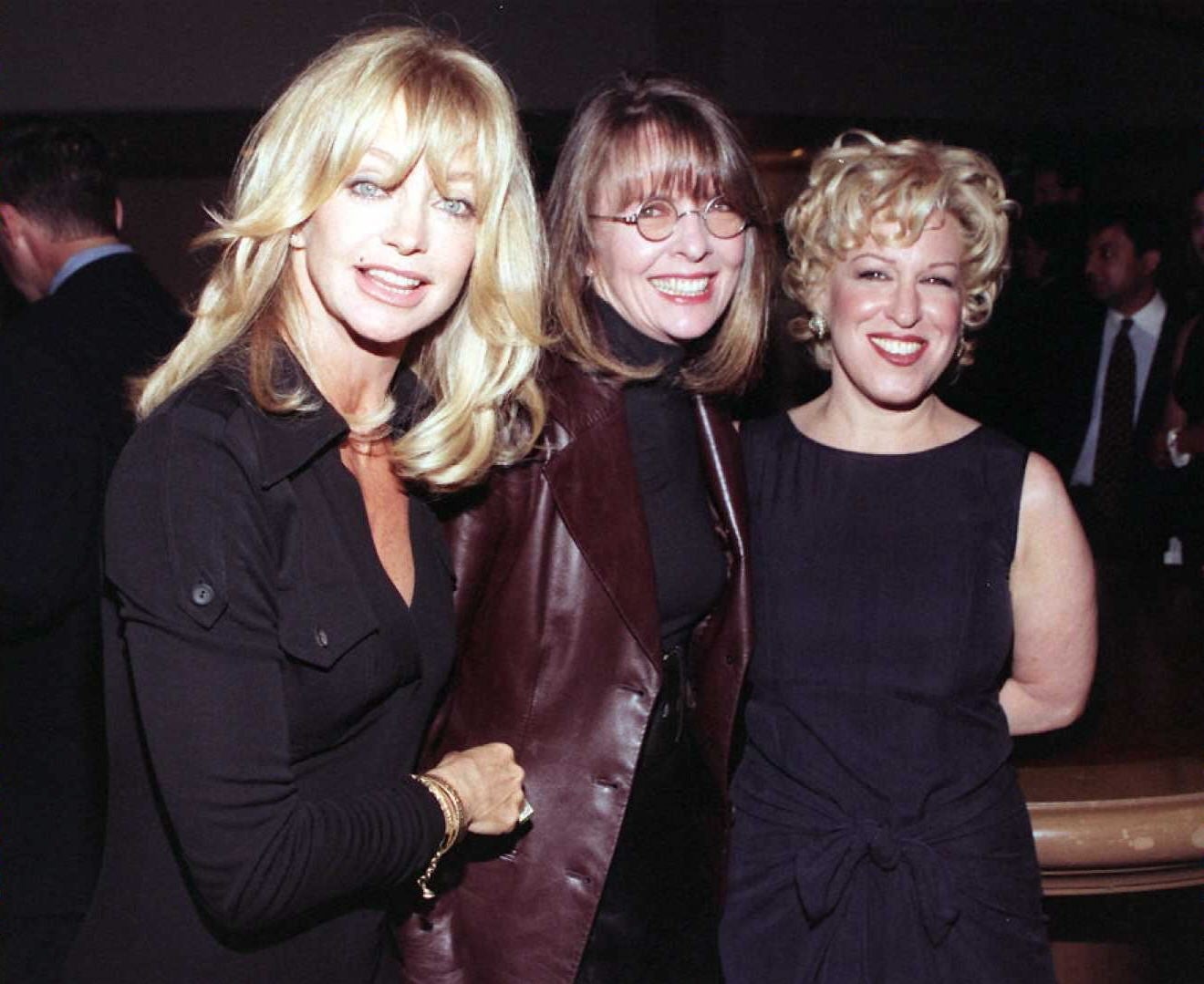 Three women smiling at an event, wearing stylish outfits; one in a black dress, another in a dark jacket, and the third in a sleeveless dress