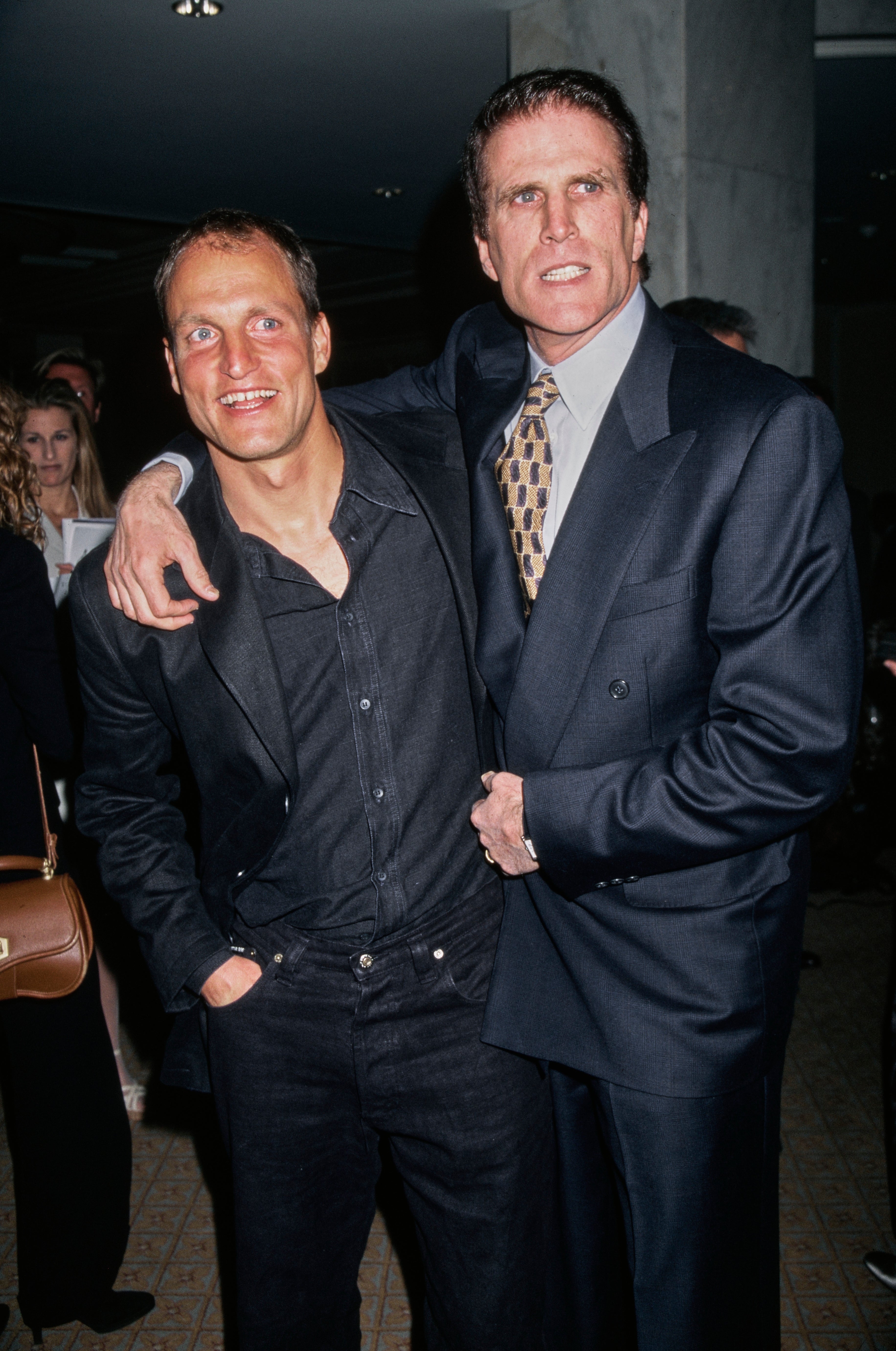 Two people in a friendly pose; one in a casual shirt and the other in a formal suit with a tie, at an event
