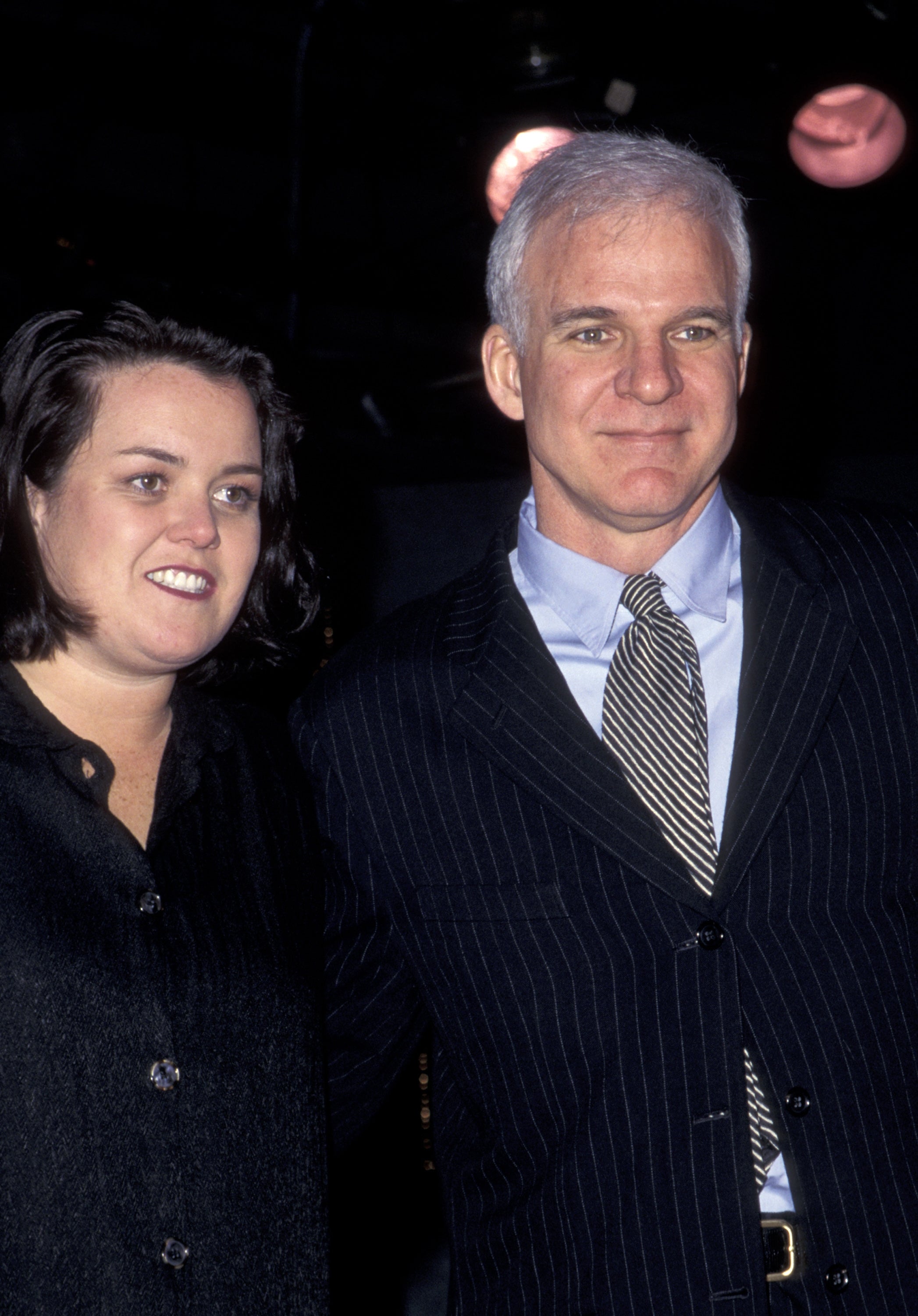 Two people smiling. One wears a dark shirt, the other in a pinstripe suit with a tie, posing together at an event