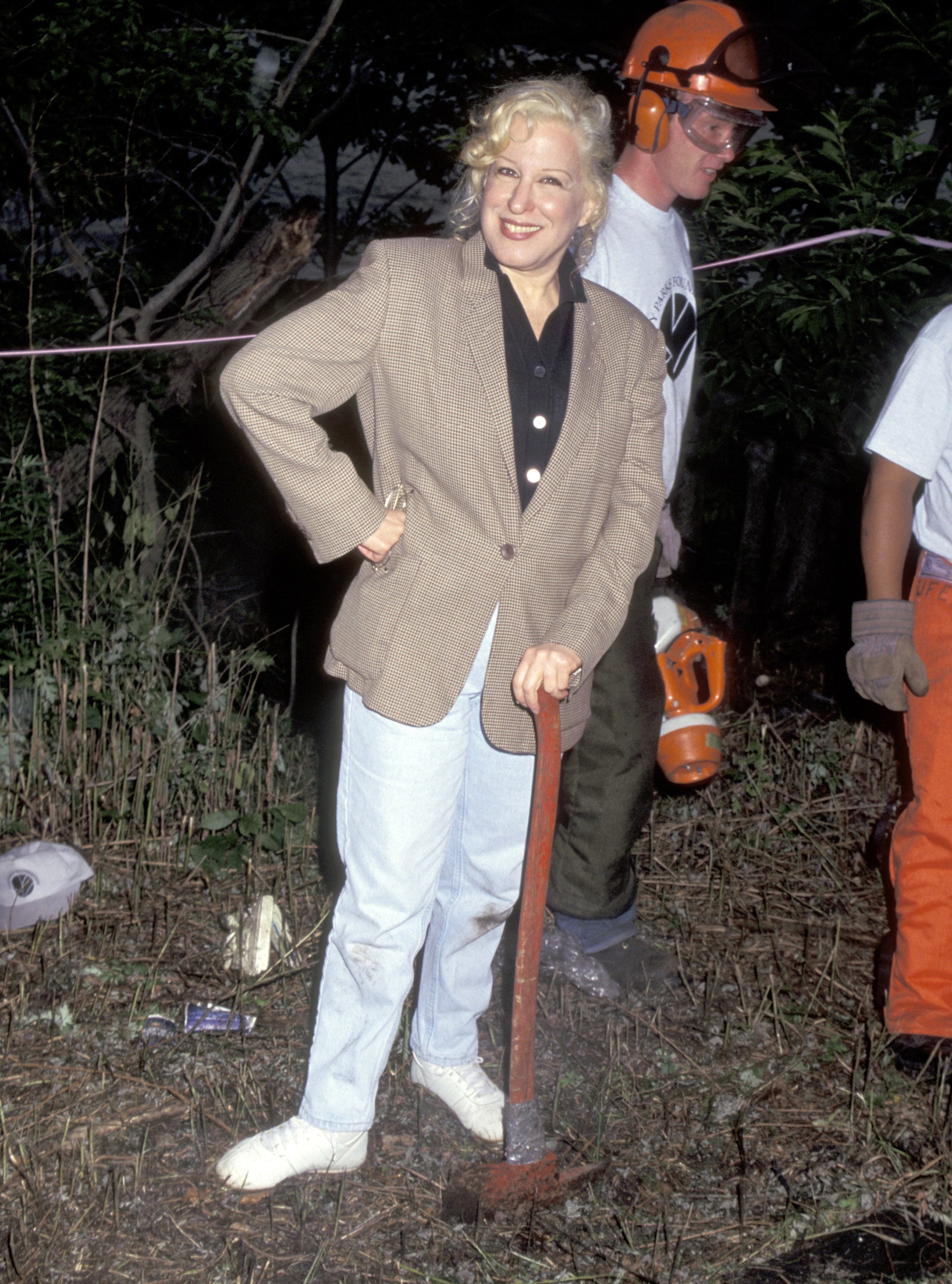 Person outdoors smiling, wearing a blazer and jeans, leaning on a shovel. In the background, two people in work attire and gear
