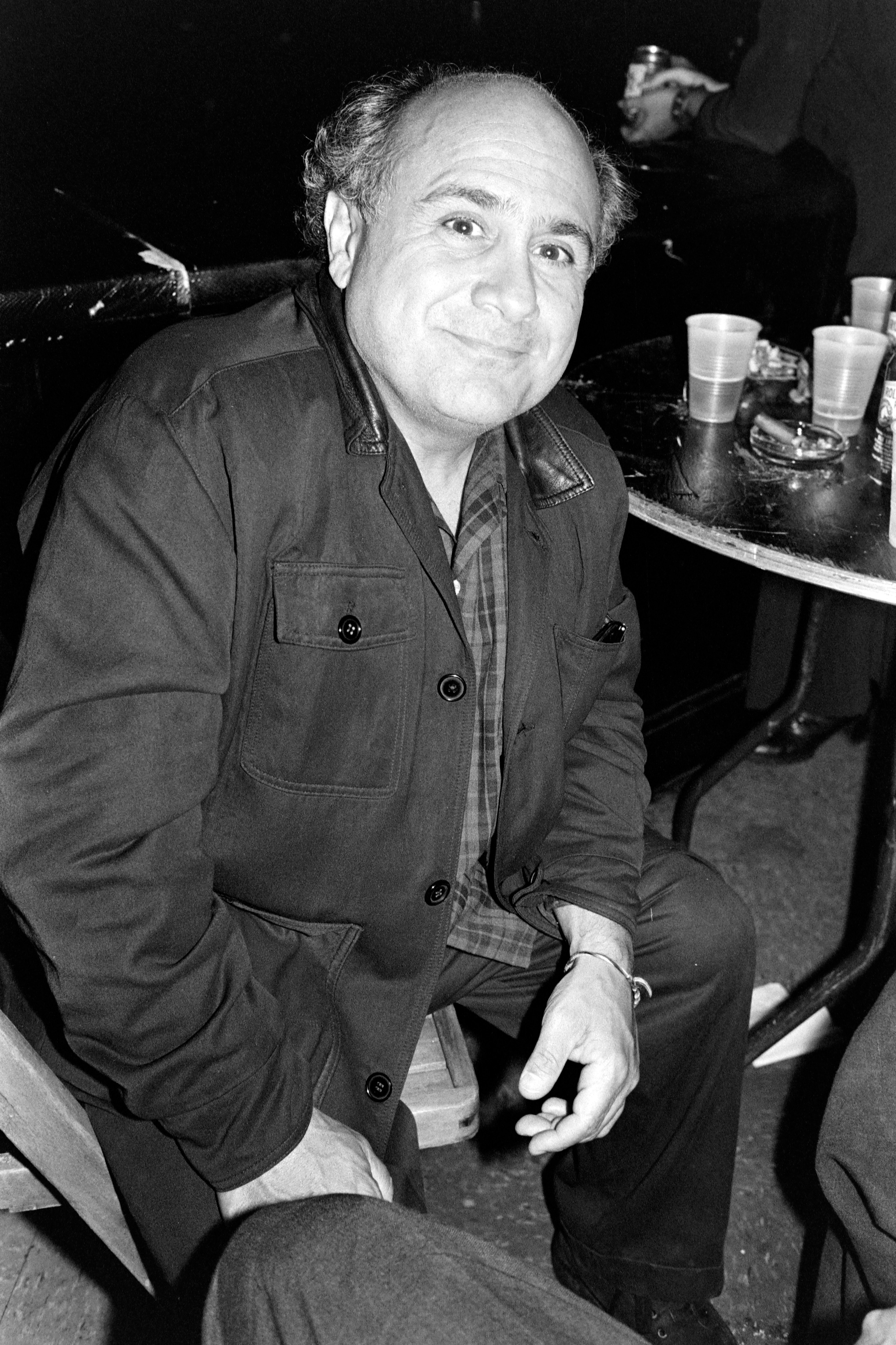 Person seated at a table with drinks, wearing a casual jacket, smiling at the camera in a candid setting