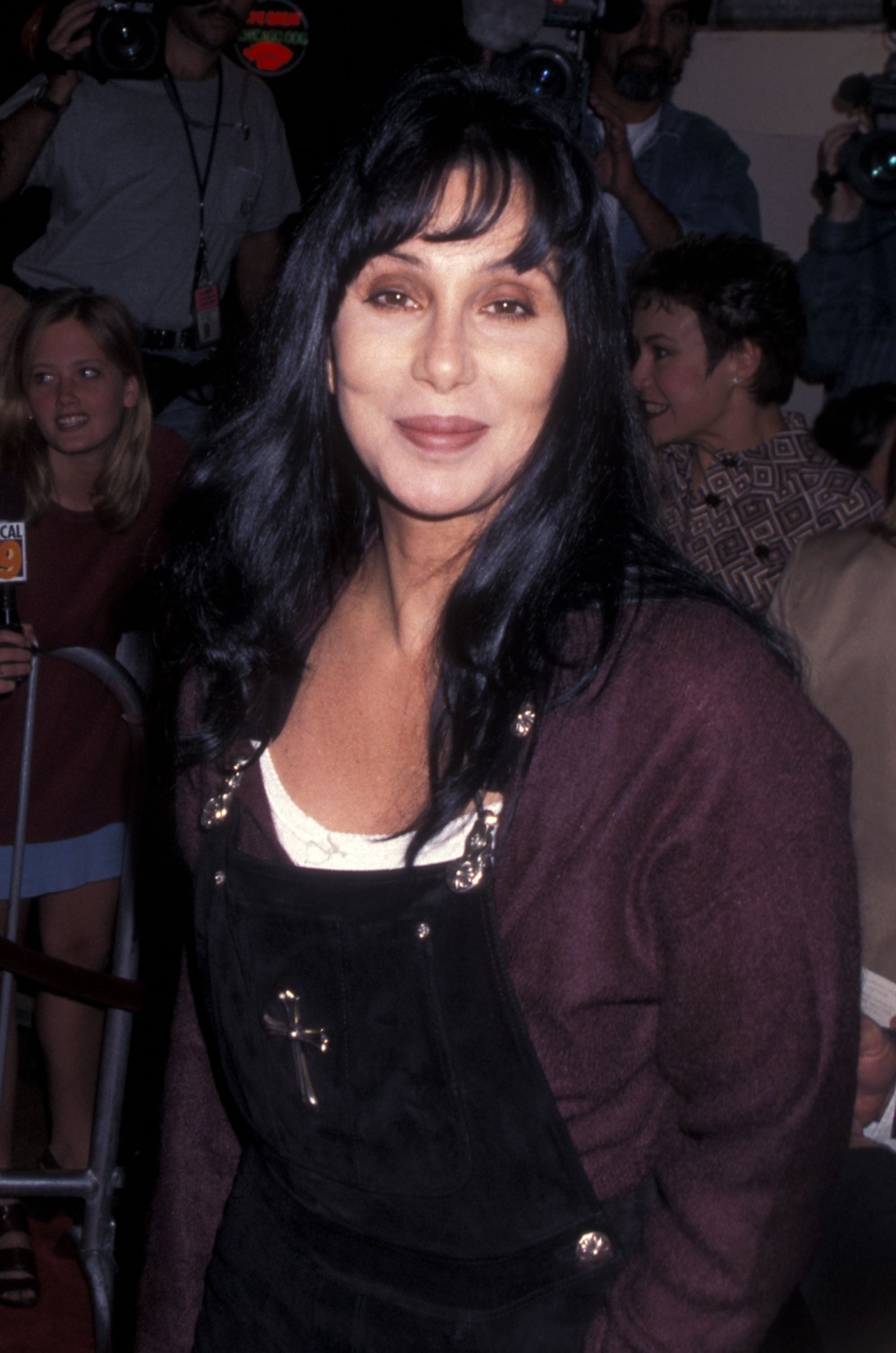 Person in layered casual wear at an event, surrounded by onlookers and photographers in the background
