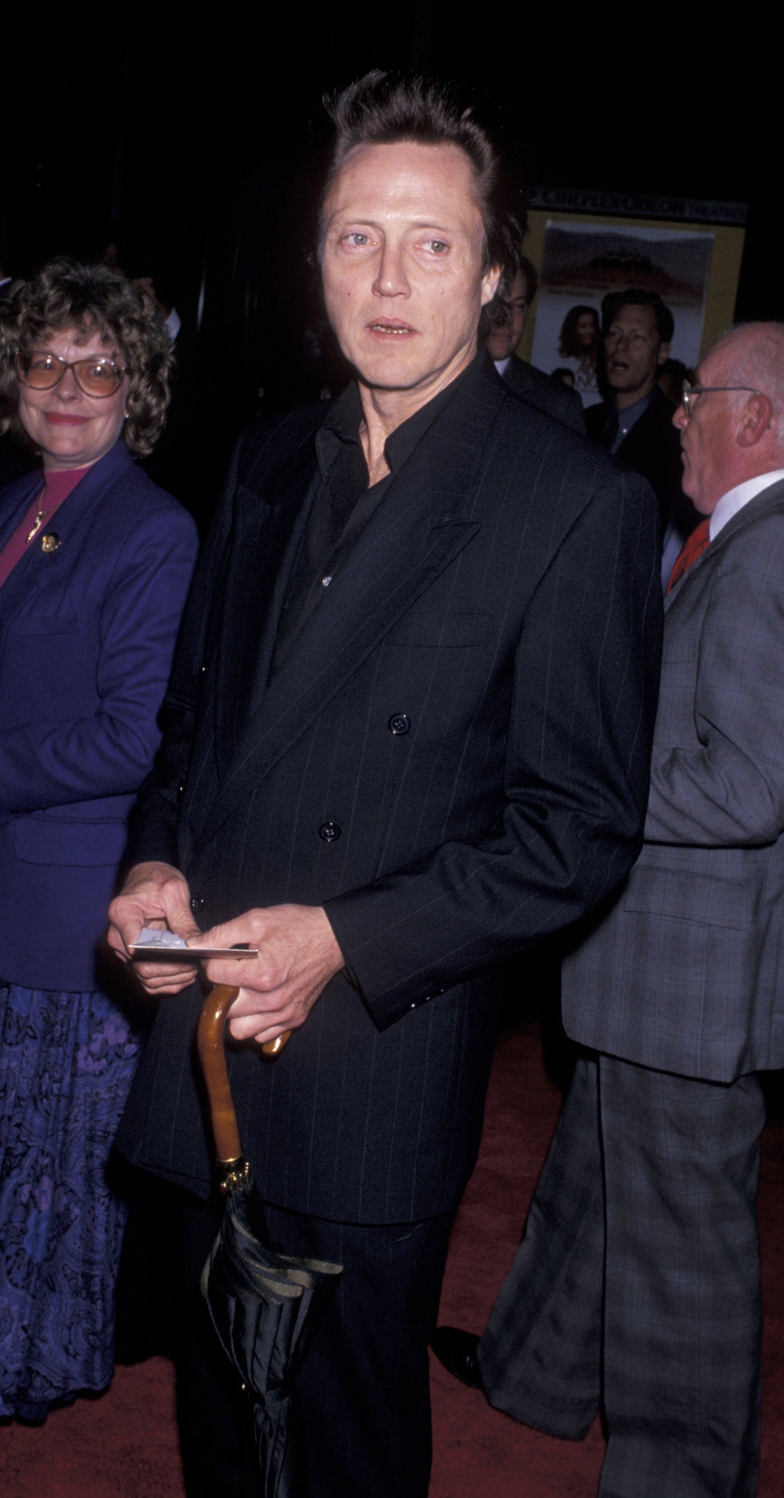 Person in a double-breasted suit holds an umbrella on a red carpet, surrounded by others in formal attire